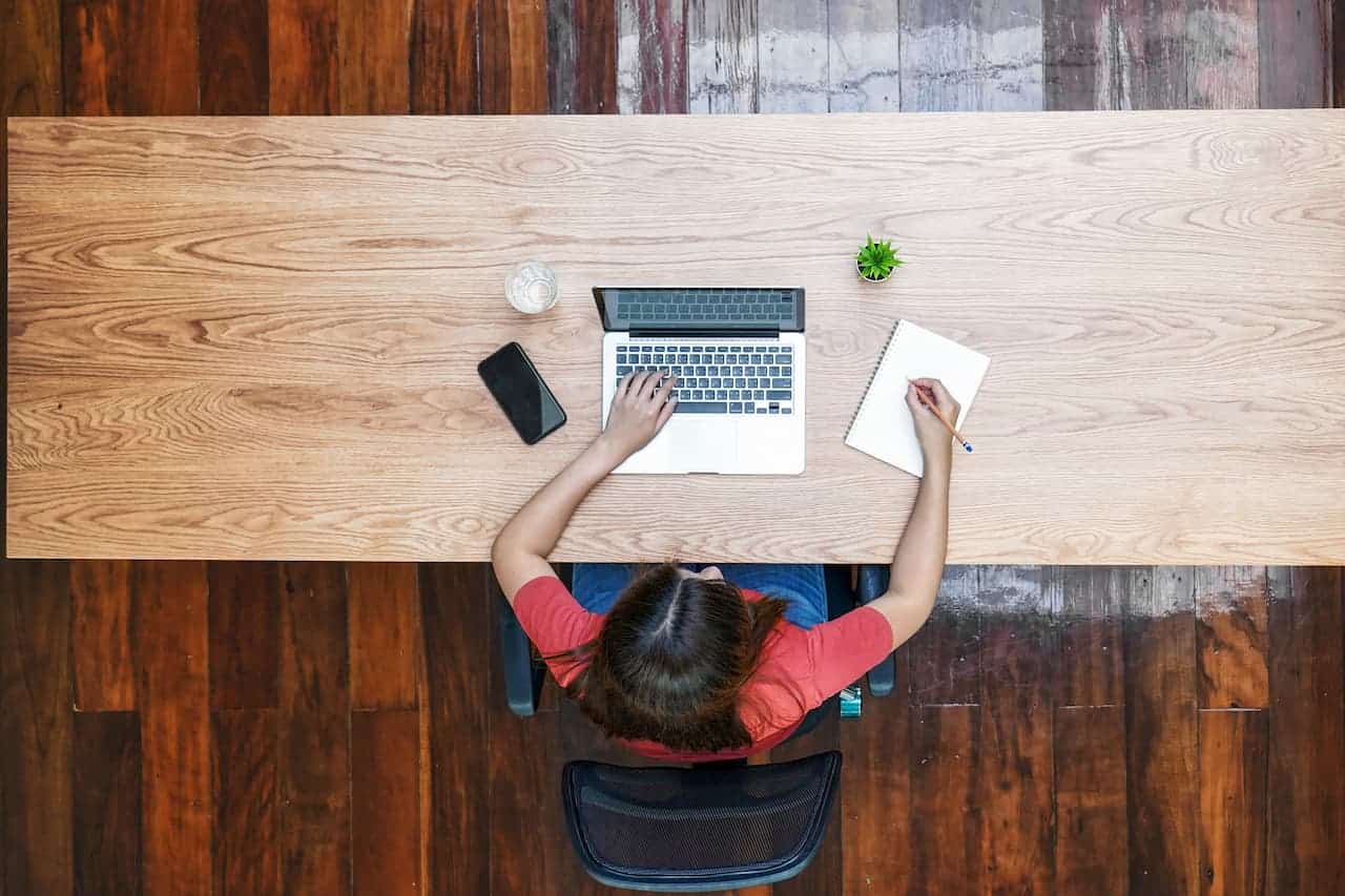A person working at a desk with a laptop, smartphone, notepad, and plant, viewed from above.