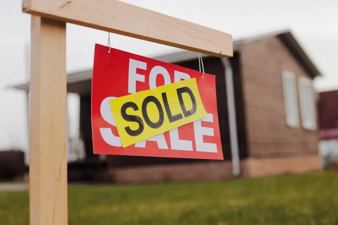 Close-up of a "Sold" sign hanging on a wooden frame in front of a house.