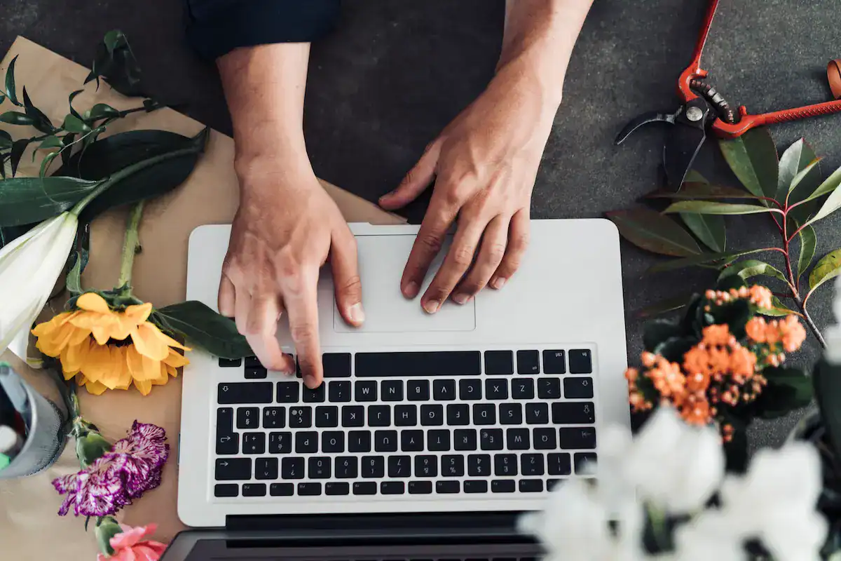 Hands typing on a laptop keyboard with flowers and gardening tools around.